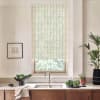 Beige plaid roller blind filters daylight over a kitchen window above a stainless sink, surrounded by a stone countertop holding a bowl of greens, potted plants, teacup and framed sketch.