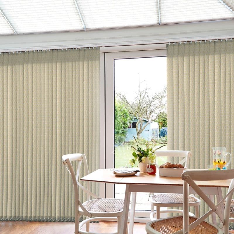 Beige patterned vertical blinds, mostly closed with a central opening, filtering daylight through patio doors; small dining table and chairs in foreground, vase and pastries on table, sunlit backyard beyond.