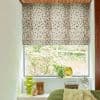 A cream Roman blind with scattered brown spots, lowered halfway and filtering daylight; over a bedroom window with plants on sill, green glass vase, vintage fan and green bedcover.