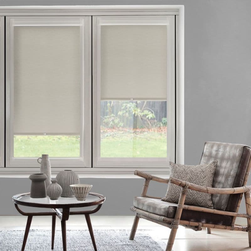 Neutral beige roller blinds, lowered halfway across two adjacent windows with visible pull tabs, filtering soft daylight; modern living room with rattan-framed leather chair, small coffee table, and garden view.