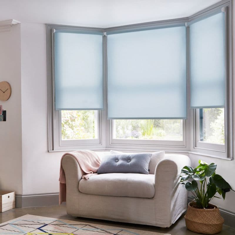 Light blue roller blinds on a three-panel bay window, lowered and semi-opaque, filtering soft daylight into a cozy living room with a beige armchair, cushion, throw and potted plant.