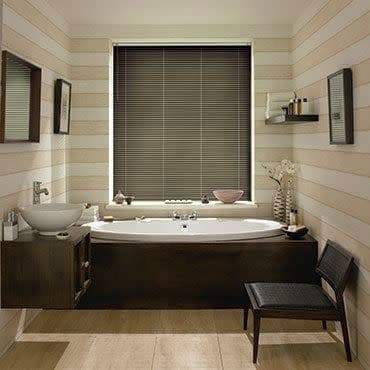 Horizontal Venetian blinds, dark gray, lowered over central window, closed and filtering light; bathroom with white bathtub in a dark wood surround, striped pale walls, vessel sink and chair.