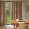 Patterned floor-to-ceiling pleated curtains, pink geometric print, drawn across a tall multi-pane window, filtering daylight into a dining room with a wooden table, chairs, and a grassy garden view.