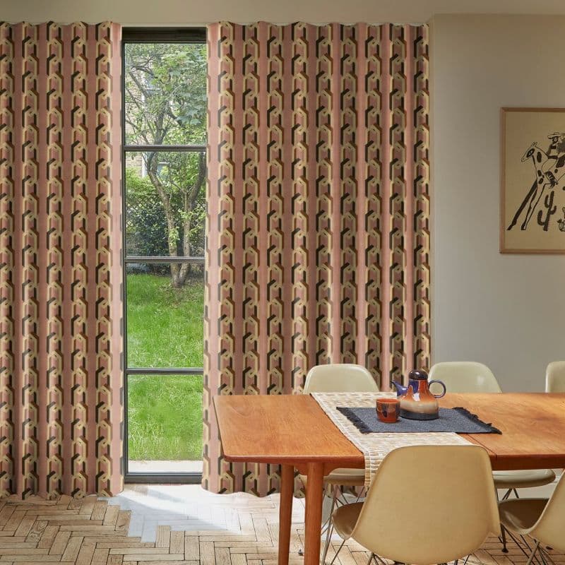 Patterned floor-to-ceiling pleated curtains, pink geometric print, drawn across a tall multi-pane window, filtering daylight into a dining room with a wooden table, chairs, and a grassy garden view.