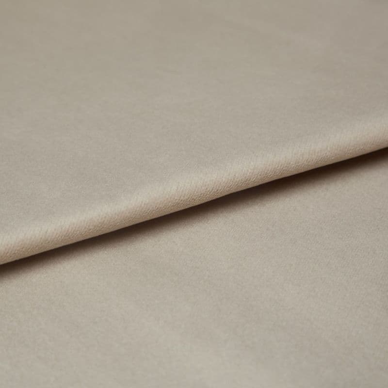 Beige velvet fabric, folded diagonally with a soft nap and slight sheen; overlapping edge creates a crease, shown in a close-up under diffuse neutral lighting on a flat surface.