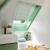 White pleated skylight blind, partially lowered over an angled roof window, filtering soft daylight; set in a bright attic bathroom with mint-green trim, white herringbone tiles, sink, tub, plant.