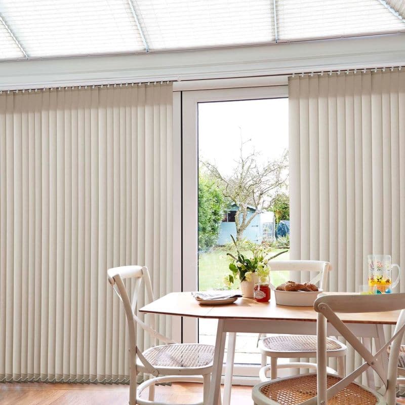 Beige vertical blinds on a sliding glass door, mostly closed with a central gap revealing the view; filtering daylight into a dining area with wooden table, chairs, and garden beyond.