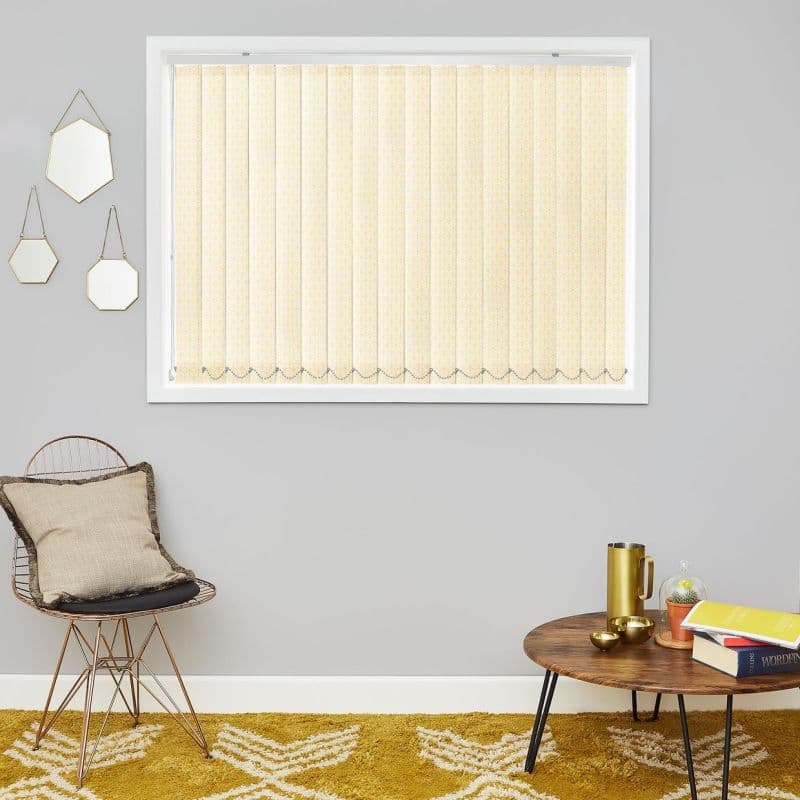 Vertical beige patterned vertical blinds, fully closed with a scalloped beaded hem, filtering daylight into a neutral living room with grey walls, a wire chair and a wooden coffee table.