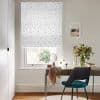 White Roman blind with small grey stars, lowered and filtering light; over a desk with teal chair, vase of flowers in a bright home office with framed seaside print.
