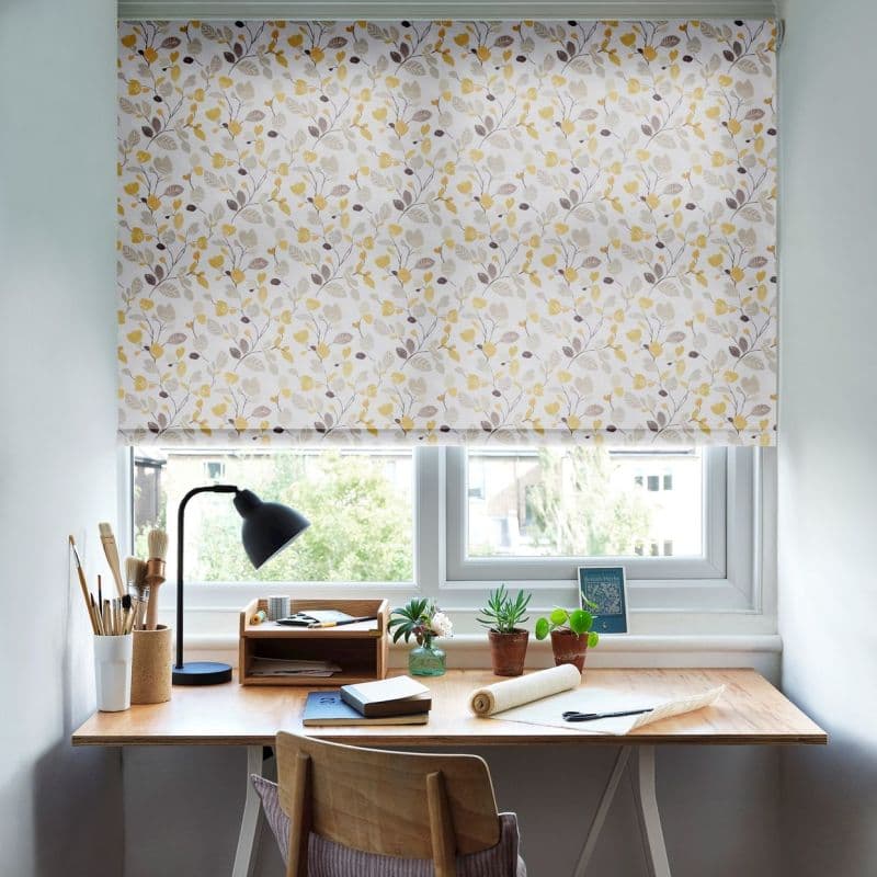 Floral roller blind with pale yellow and beige leaf pattern, fully lowered over a double window, softly filtering daylight; compact desk beneath holds a lamp, brushes, notebooks, and potted succulents.