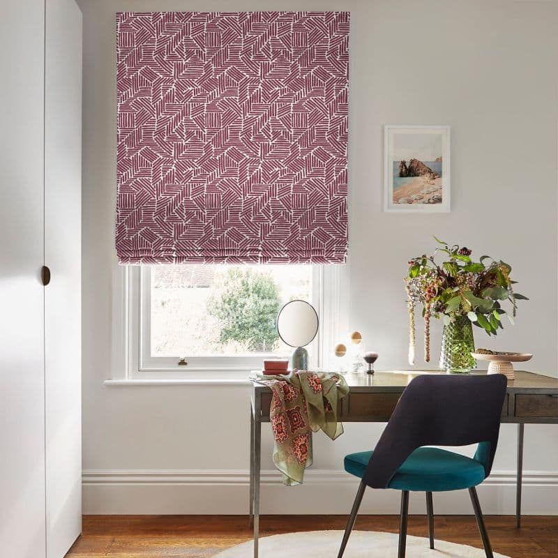 Burgundy-and-white geometric Roman blind, lowered with a soft fold at the bottom, filtering daylight. Small home office: wooden desk with mirror, teal chair, vase of flowers, pale walls, garden view.