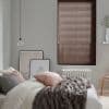 Brown horizontal slatted blinds, lowered and closed, slightly filtering daylight; bedroom setting with grey walls, white bed topped by pink and grey pillows and a grey faux-fur throw, radiator below.