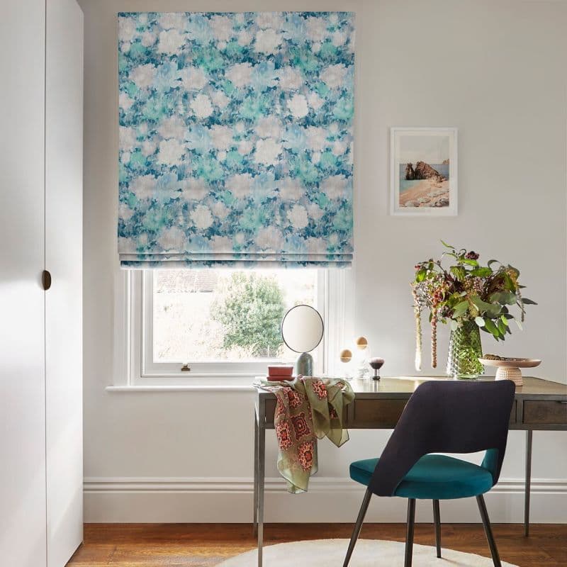 Roman blind with blue watercolor pattern, lowered and folded, filtering daylight through the window; above a small desk with teal chair, vase of flowers and framed print, facing a garden.