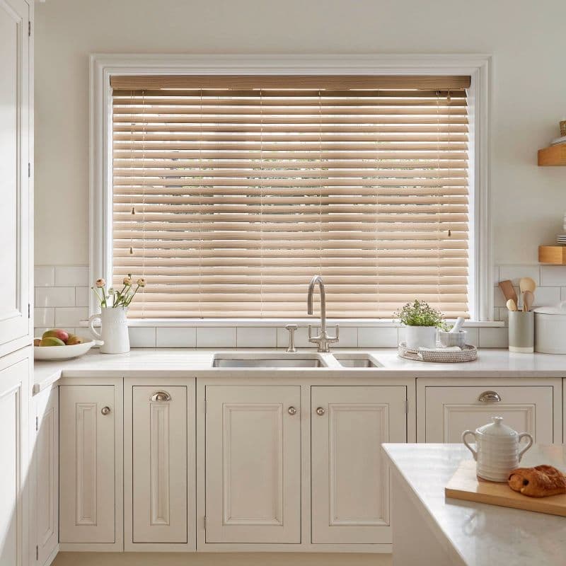 Wooden horizontal blinds (light oak slats, pull cords) mostly closed over a window, filtering daylight into a bright neutral kitchen with a double sink, marble counters and potted herbs.