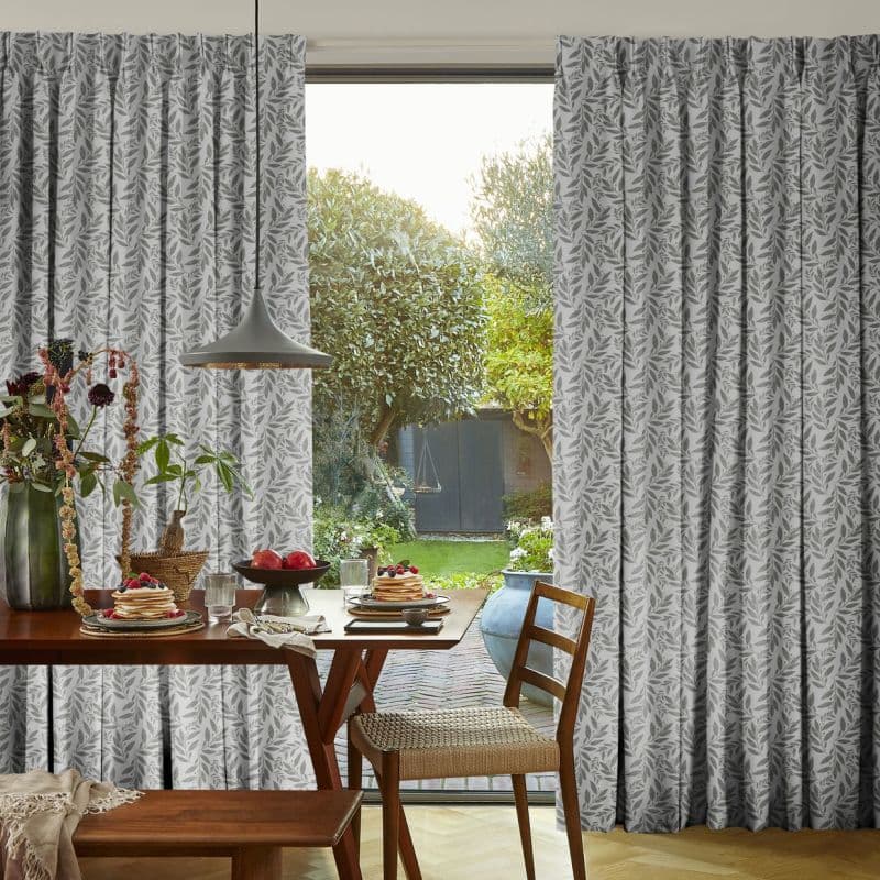 Gray leaf-patterned pleated curtains framing a sliding glass door, parted at center and softly filtering daylight; a breakfast table with pancakes and pendant lamp faces a sunlit garden and shed.