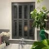 Dark grey four-panel interior louvered shutters covering window, panels closed with slats angled to filter light; set above a bathtub with chrome faucet, grey subway tiles, orchids and houseplants nearby.