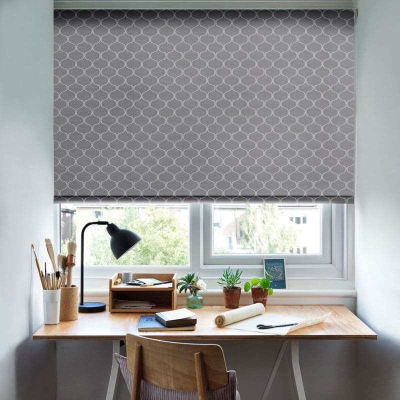 A grey roller blind with white trellis pattern lowered over a double window; it filters daylight. Below, a tidy wooden desk holds a lamp, brushes, potted plants and notebooks.
