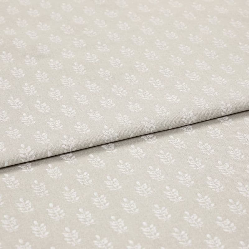 Beige cotton fabric with small white leaf motif, folded once to reveal overlapping layers; lying flat on a neutral surface under soft, even studio lighting for a close-up textile display.