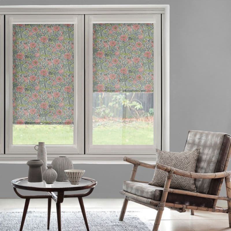 Floral patterned roller blinds (pink poppy motif) on dual windows — left fully lowered, right half-raised — filtering daylight into a grey-walled sitting area with rattan chair and table.