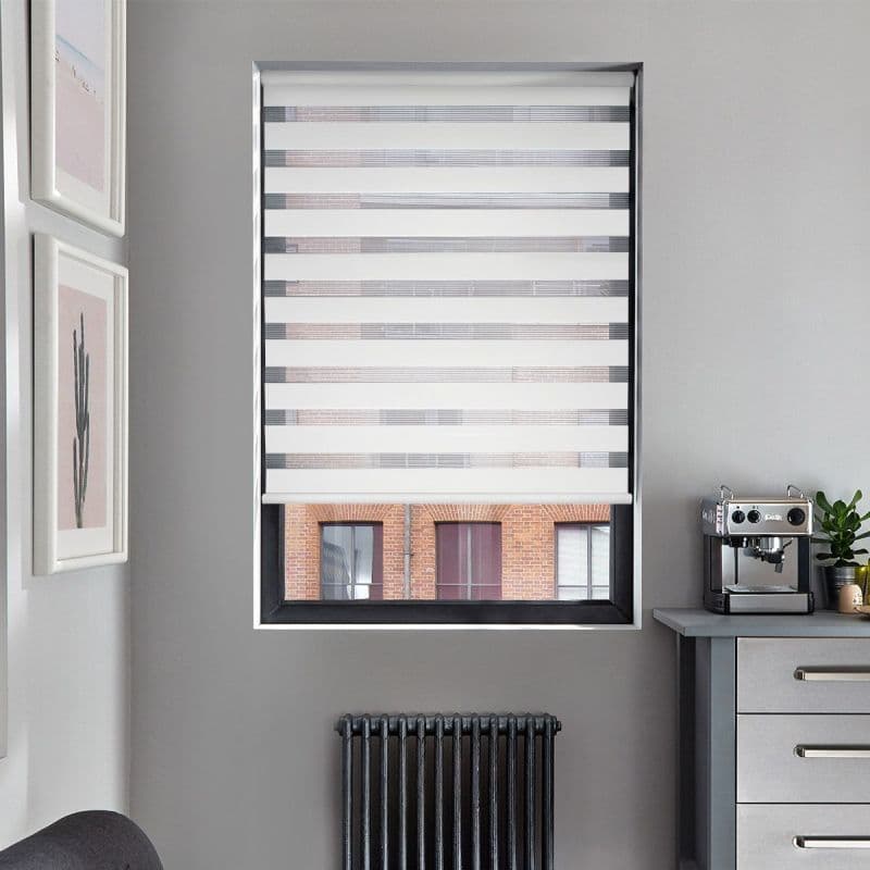Zebra-style roller blind, lowered with alternating opaque and sheer horizontal bands, partially revealing bottom pane; filtering daylight in gray kitchen corner with coffee machine, radiator and framed prints.