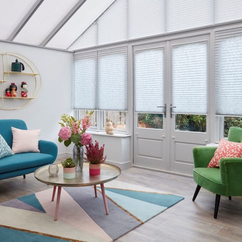 White pleated cellular blinds cover tall conservatory windows and glazed doors, lowered to mid-height, softly filtering daylight; bright sunroom with teal sofa, green armchair, pastel rug, coffee table with flowers.
