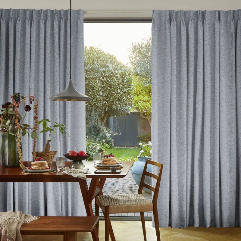 Gray pleated floor-length curtains, parted center to reveal glass door, soft textured fabric. They frame a sunlit dining nook with wooden table, pendant lamp, and a green backyard with trees.