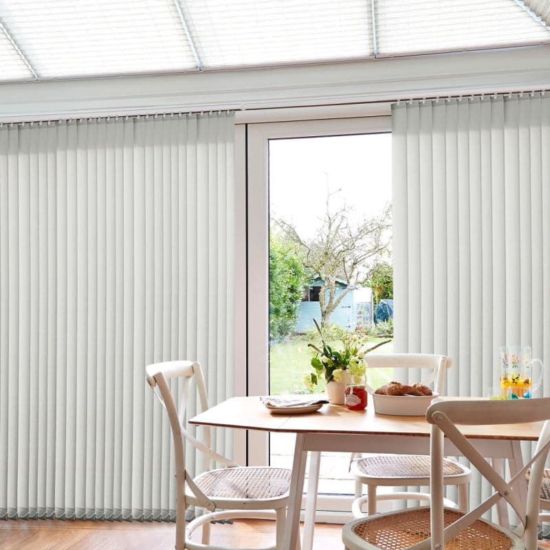 Cream vertical blinds, floor-to-ceiling panels split to reveal a central glazed patio door, partially open; filtering daylight into a dining area with wooden table, wicker chairs and a garden view.
