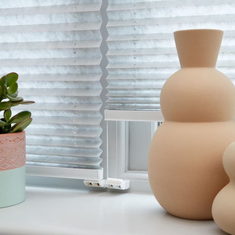 White pleated cellular blinds lowered with a central zigzag seam and visible cord beads, filtering soft daylight on a windowsill beside a pastel succulent pot and two rounded beige vases.