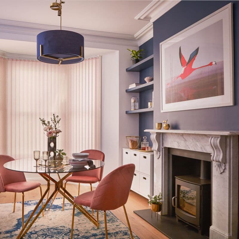 Pale-pink vertical blinds covering a bay window, fully closed and softly diffusing daylight; a dining room with round glass table, blush velvet chairs, navy pendant, marble fireplace and flamingo artwork.