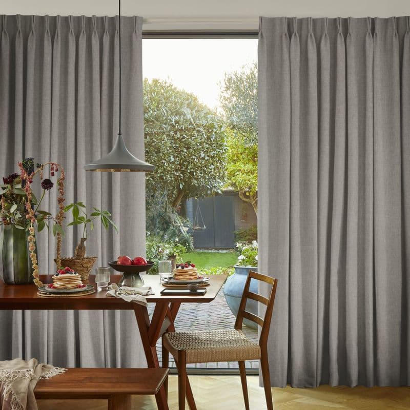 Gray floor-to-ceiling pleated curtains, slightly parted in the center with soft folds, revealing a sunlit backyard; dining table with breakfast settings, pendant lamp and wooden chair in foreground.