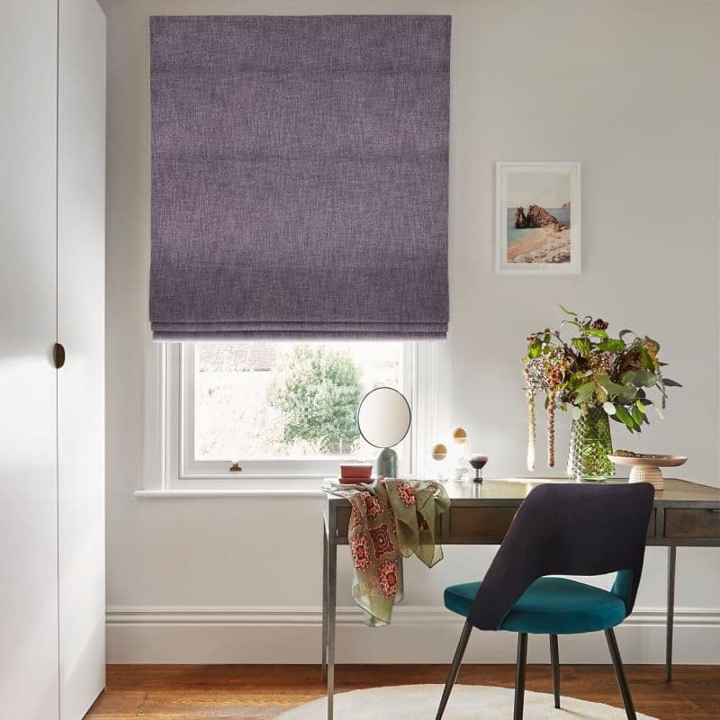 A textured purple Roman blind, fully lowered with horizontal folds, filters daylight over a sash window; a wooden desk, teal chair, vase of flowers and framed seaside print sit below.