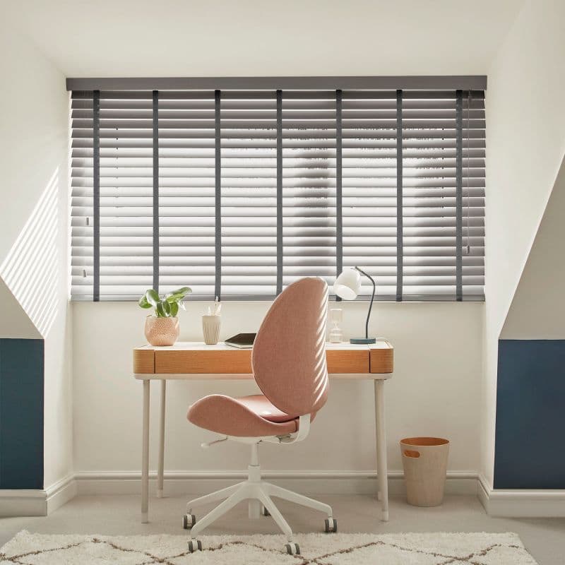 Grey horizontal slatted blinds covering a wide dormer window, slightly tilted to filter sunlight; above a desk in a bright attic study with a pink swivel chair, lamp and plant.