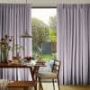 Floor-length pleated lavender curtains, drawn slightly apart to reveal a central opening, softly filtering daylight; dining table with pancakes and pendant lamp in foreground, backyard garden visible beyond.