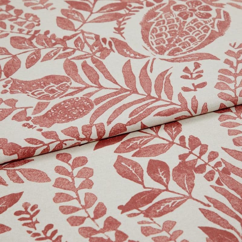 Cream fabric printed with terracotta botanical and pomegranate motifs, folded and overlapped to show pattern repeat, close-up on a flat surface under soft, even studio lighting.