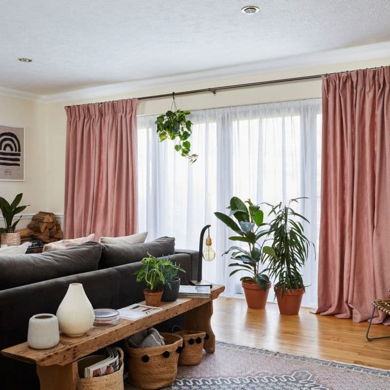 Pink gathered drapes with white sheer panels on a rod, filtering daylight and framing a sliding glass door in a bright living room with dark sofa, bench, and potted plants.
