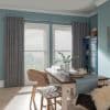Patterned drapes on a rod flank two tall windows; lowered white horizontal blinds filter daylight in a sunlit teal dining room with wooden table, chairs, table runner and a blue cabinet.