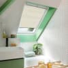 Cream pleated skylight blind, lowered halfway and filtering daylight, in a bright sloped-ceiling bathroom with green trim, white herringbone tiles, sink, bathtub and potted plant.
