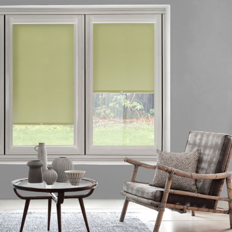 Light-green roller blinds on two adjacent windows, lowered with small bottom gaps, filtering daylight; in a grey-walled living room overlooking a grassy yard, with a rattan armchair and coffee table.