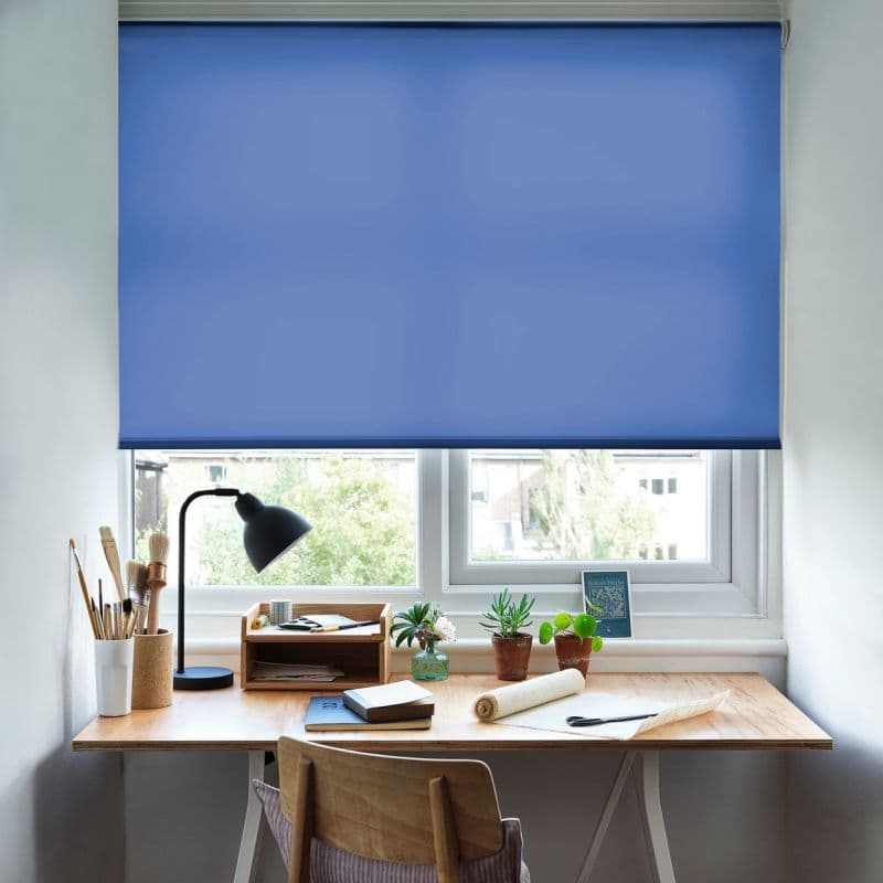 Blue roller blind, lowered across a double window with smooth fabric, filtering daylight; a wooden desk beneath holds a lamp, paintbrushes, notebooks and potted succulents, overlooking a suburban view.