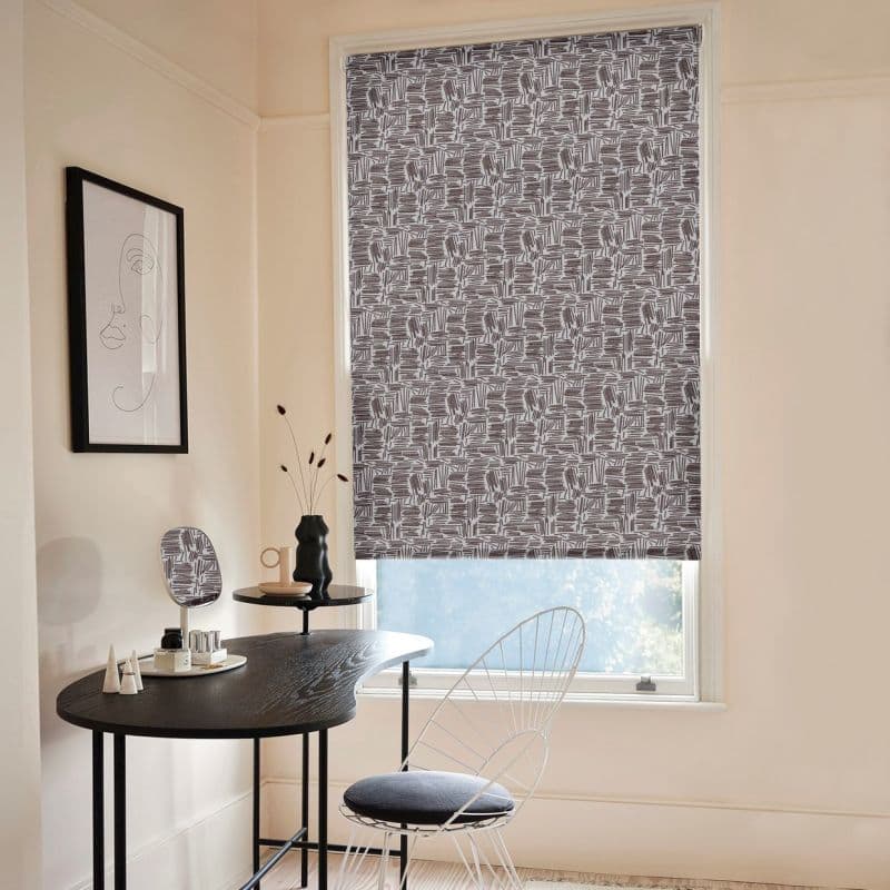 Patterned roller blind, grey-and-white geometric scribble print, fully lowered across the window; filtering daylight into a pale-walled home office nook with a black desk, wire chair, and minimalist decor.