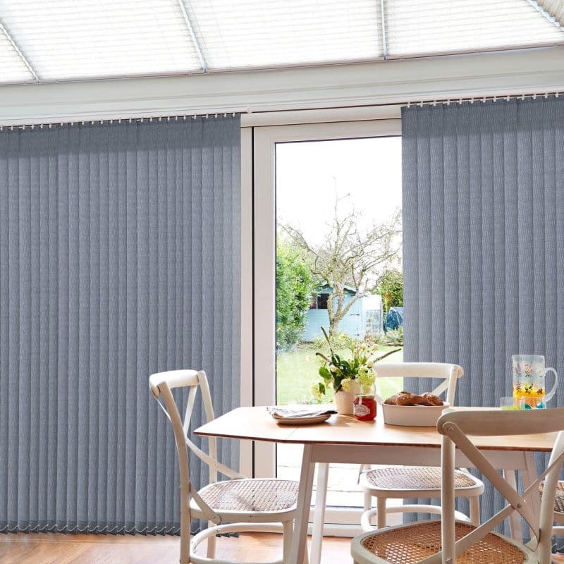 Grey vertical blinds, lowered and mostly closed with a central gap over a glass patio door, filtering light; bright dining area with table, white chairs and a garden view.