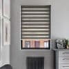 Zebra roller blind, fully lowered with alternating opaque and sheer horizontal bands; filtering daylight and revealing glimpses of a brick building, in a gray nook with coffee machine and radiator.