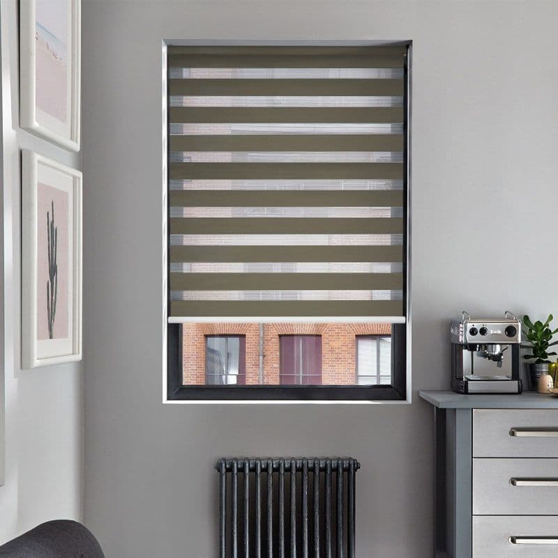 Zebra roller blind, fully lowered with alternating opaque and sheer horizontal bands; filtering daylight and revealing glimpses of a brick building, in a gray nook with coffee machine and radiator.