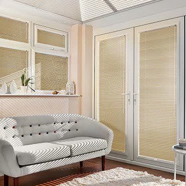 Beige horizontal blinds cover French doors and windows, fully closed and filtering sunlight, casting striped shadows across a cream tufted sofa, windowsill plants, wooden floor and white rug.