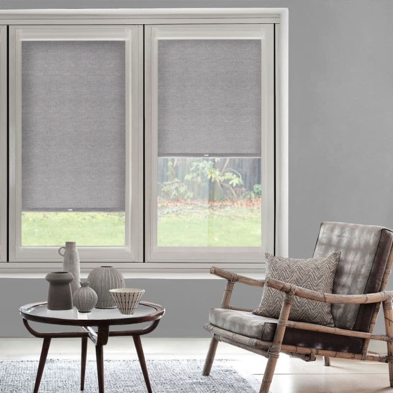 Grey fabric roller blinds, partially lowered across two adjacent windows, filtering daylight; living room with round coffee table holding ceramic vases and a rattan leather armchair, backyard lawn visible.