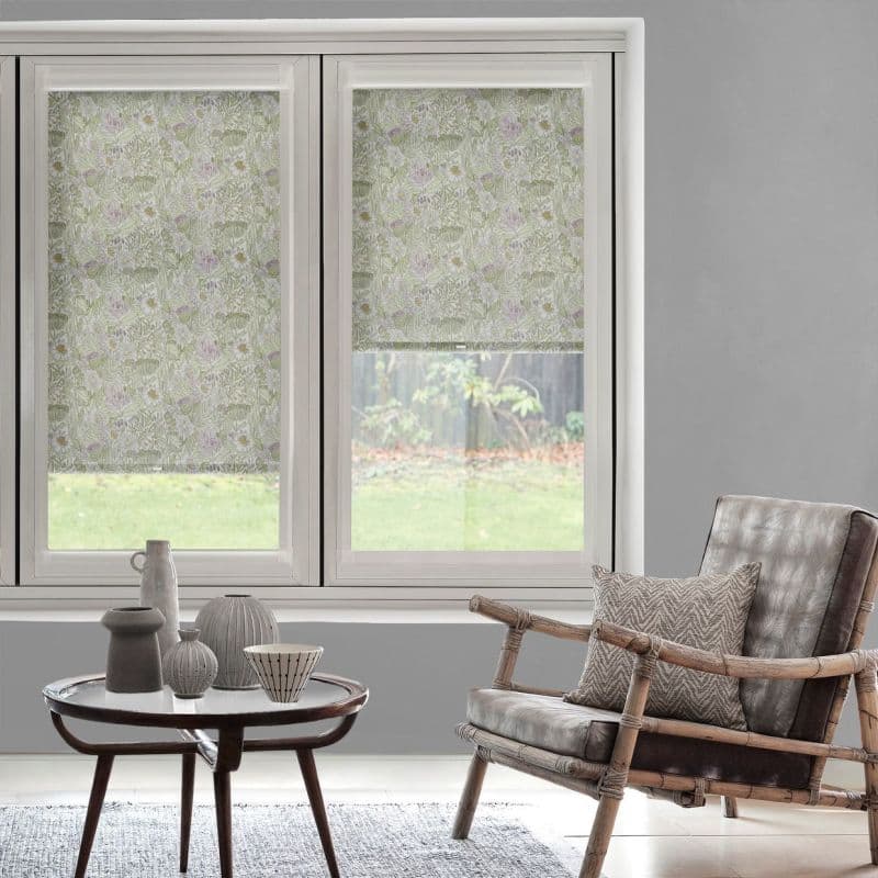 Floral-patterned roller blinds covering two adjacent windows, right blind slightly raised; filtering soft daylight into a neutral-toned living room with rattan armchair, low table, and grassy backyard view.
