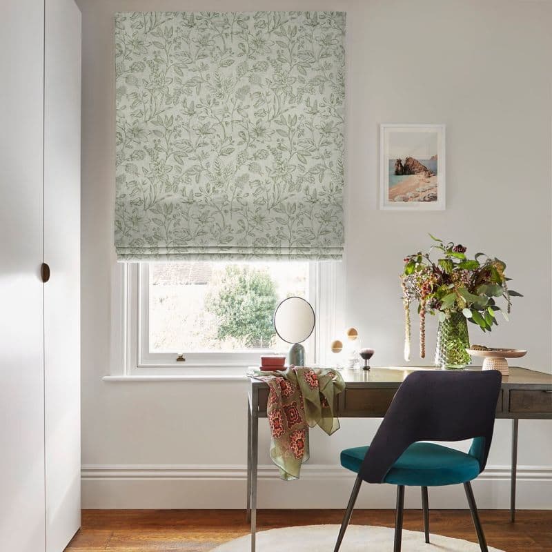 Floral-patterned Roman blind (pale green botanical print) lowered over a window, filtering soft daylight — home study desk beneath with mirror, vase of flowers, teal chair and neutral walls.
