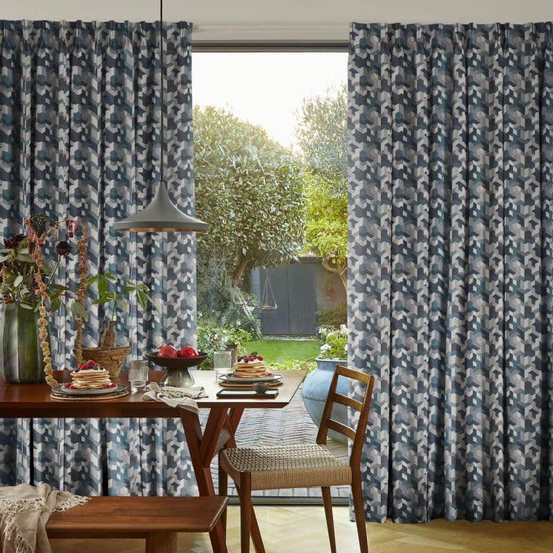 Full-length pleated geometric blue-gray curtains, drawn to sides and framing a central sliding glass door, filtering soft daylight; dining room with wooden table set for breakfast, pendant lamp, leafy backyard.