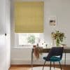 Pale mustard fabric Roman blind, fully lowered, filtering daylight through a small lower gap; above a wooden desk with teal chair, vase of flowers, framed print and garden view.