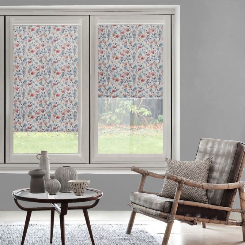 Floral-patterned roller blinds, lowered over two side-by-side windows (right slightly raised), filtering daylight; modern living room with grey walls, wicker armchair and cushion, small coffee table holding ceramics.